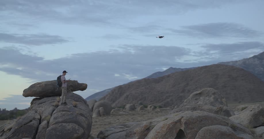 A drone pilot stands on a rocky outcrop in a vast desert landscape, operating his drone as it flies around him with majestic mountains in the background