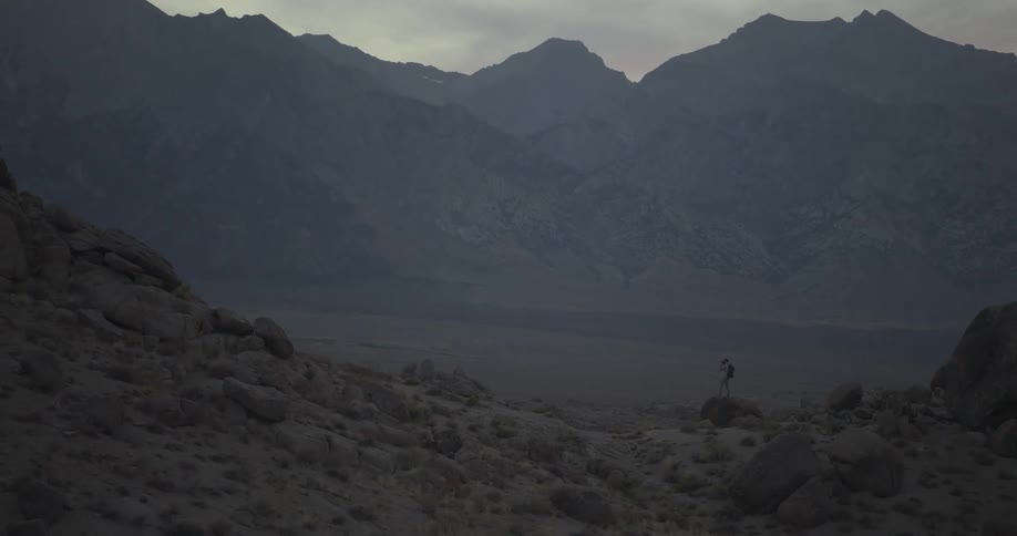 A solitary hiker stands on a rocky hill, dwarfed by the immense and moody mountain range at dusk