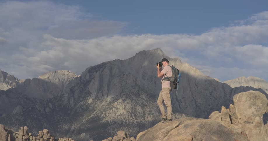 An adventurous photographer stands on a large boulder taking pictures of the majestic mountain range in a vast, rocky landscape