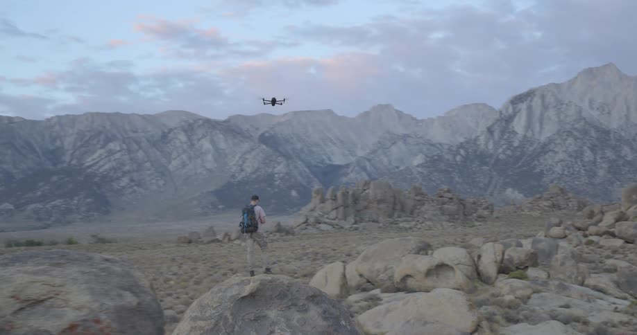 A hiker stands on a large boulder and flies his drone through a vast, rocky landscape with towering mountains in the background