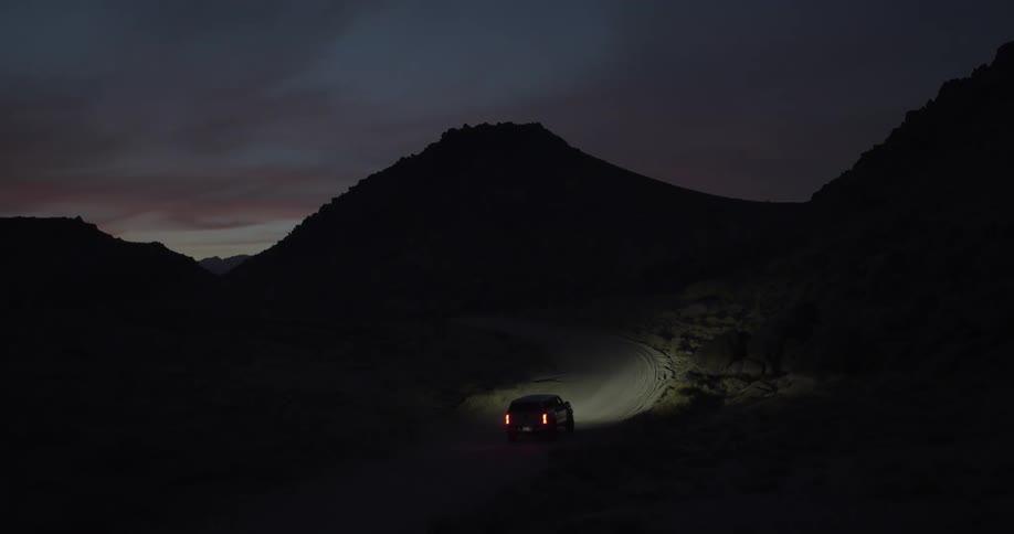 An aerial view follows a lone SUV as it navigates a winding dirt road through a dark mountain pass at dusk
