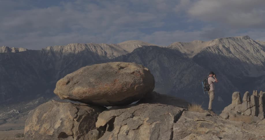 A photographer stands on a rocky outcrop in a vast desert, using binoculars to view the majestic Sierra Nevada mountains before walking out of frame