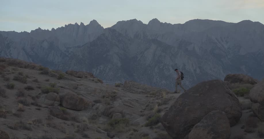 Slow motion shot of a hiker with a backpack trekking across a desolate, rocky landscape with mountains in the distance at twilight