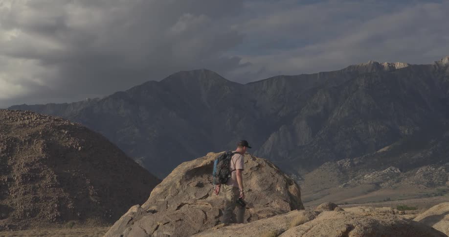 A male hiker wearing a backpack carefully navigates a rocky desert trail in slow motion, with a dramatic mountain range in the background