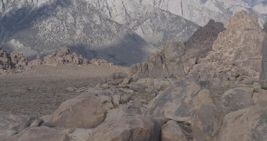 A lone hiker with a backpack walks through a vast, rocky desert landscape at the base of a towering mountain range