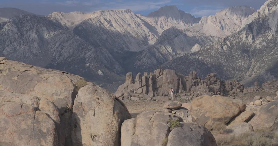 A hiker with a backpack stands on a large boulder, admiring the view of the majestic Sierra Nevada mountains in the Alabama Hills