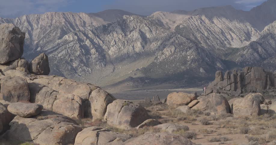 A slow pan reveals a lone hiker standing amidst the rocky landscape of the Alabama Hills with the majestic Sierra Nevada mountains in the background
