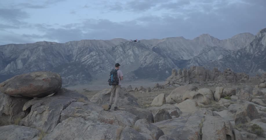 A male hiker with a backpack pilots a drone from a large boulder in a vast mountain landscape at dusk