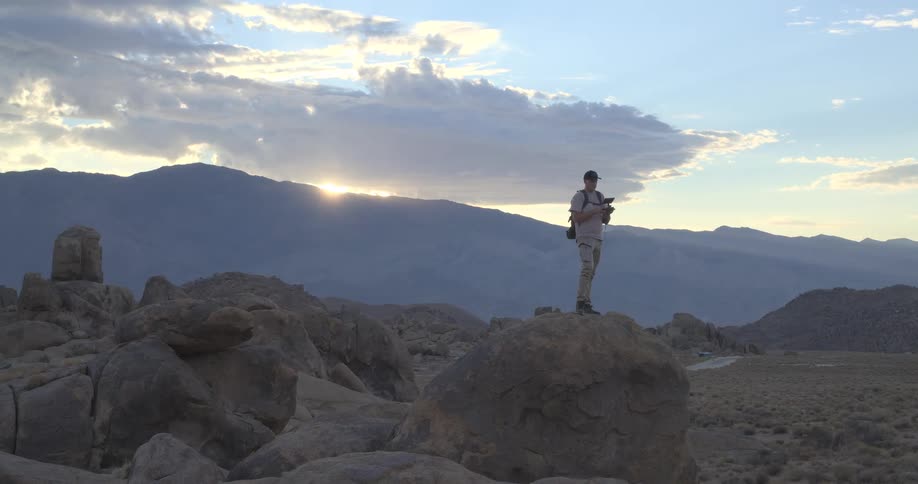 A drone pilot stands atop a boulder in the Alabama Hills, operating his remote against a stunning mountain sunset