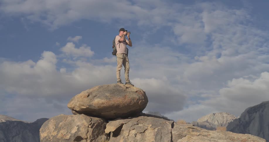 A male photographer with a backpack stands atop a large boulder, capturing photos of the majestic mountain landscape