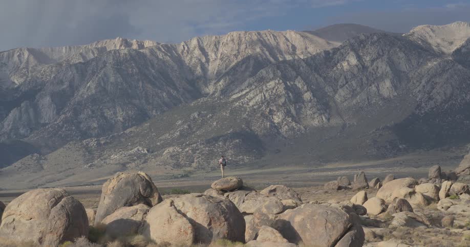 A lone hiker with a backpack stands on a large boulder, taking in the epic view of the Sierra Nevada mountain range