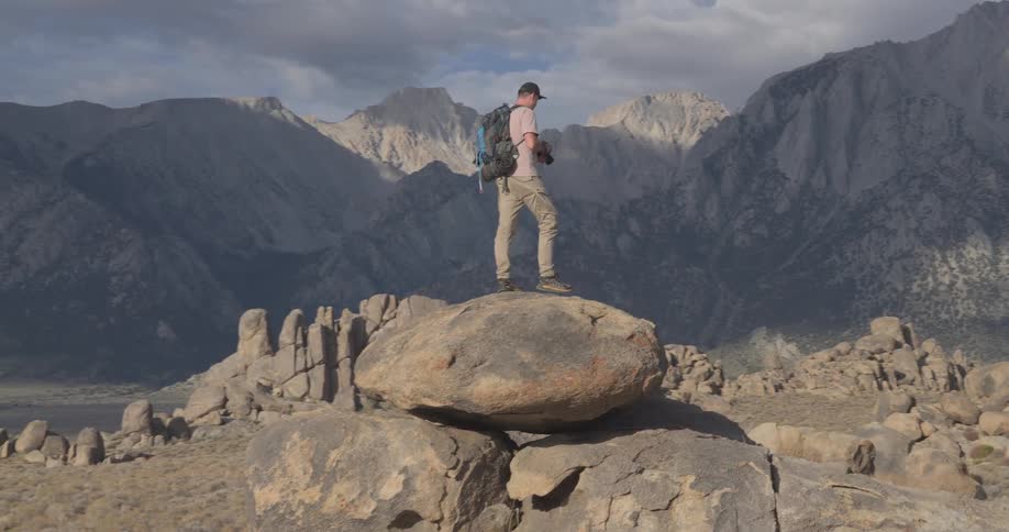 A male hiker with a backpack stands on a large boulder in a rocky landscape and takes photos of the majestic mountain range