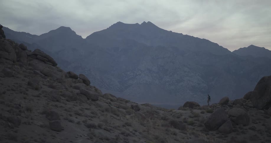 A lone hiker with a backpack walks through a vast, rocky mountain landscape at dusk