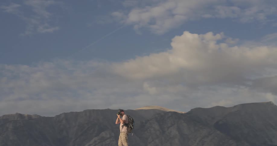 A slow pan reveals a photographer with a backpack standing atop a massive boulder, taking pictures of the epic mountain landscape