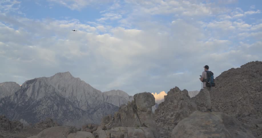 A hiker launches a drone in a vast, rocky landscape, with the camera following its flight high into the sky above majestic mountains
