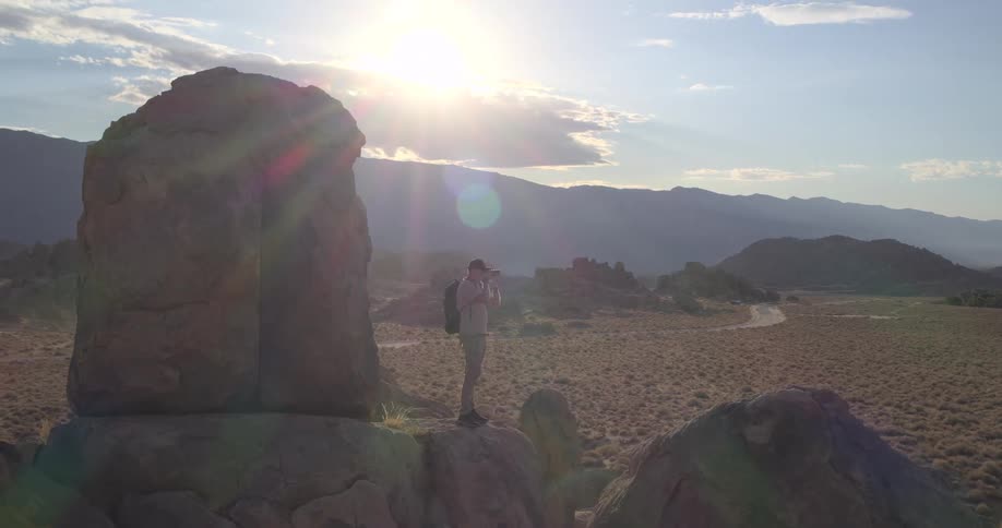 A lone photographer hikes onto a large rock formation in the desert to capture the stunning mountain landscape as the sun sets
