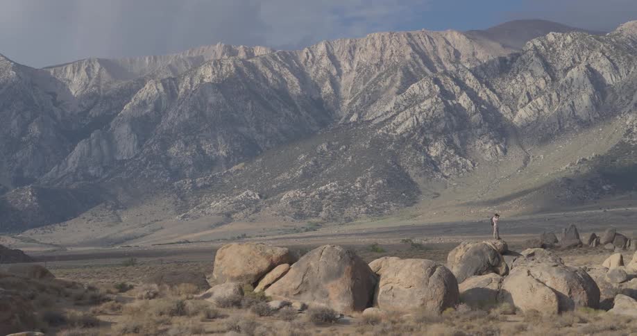 A lone explorer stands atop a large boulder, dwarfed by the immense mountain range in a vast desert landscape