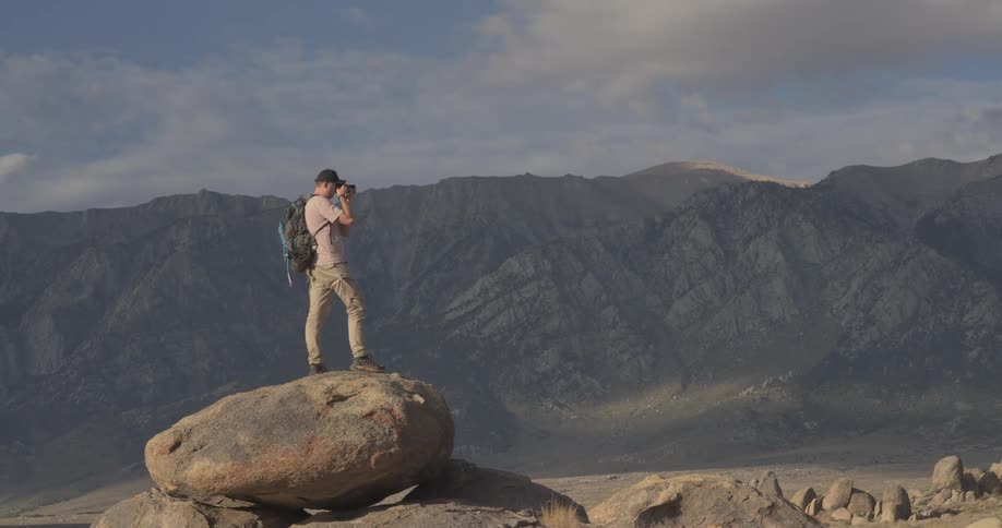 A male photographer with a backpack stands on a large boulder, capturing photos of the expansive mountain range in front of him