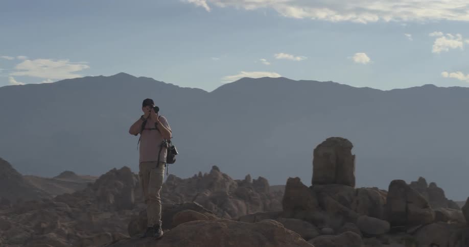 A photographer with a backpack stands atop a large rock in the desert, capturing photos of the expansive mountain scenery as the camera slowly orbits
