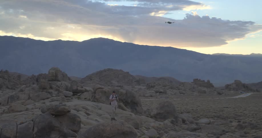 A man stands on a large rock in a vast desert landscape operating a drone that flies over the mountains at sunset