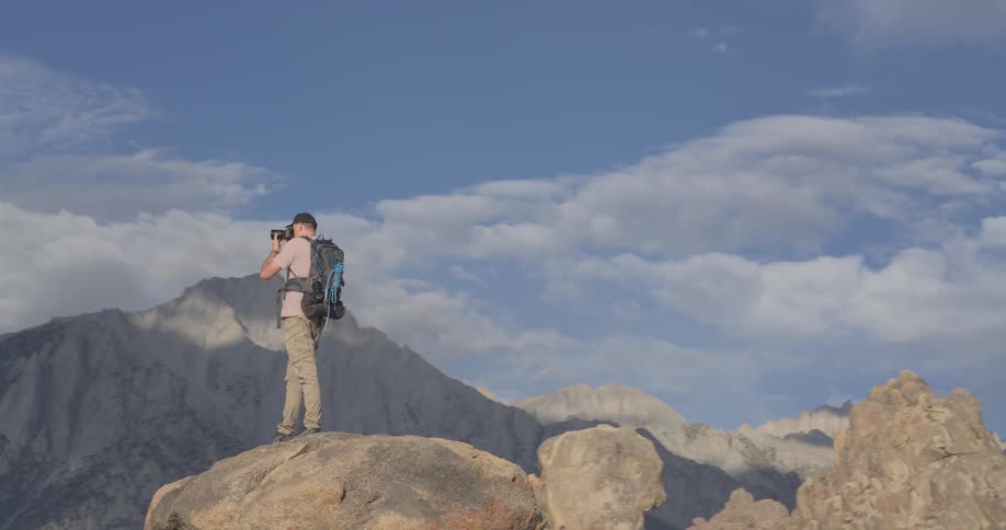 An adventurous photographer with a large backpack stands atop a giant boulder, capturing the breathtaking view of the majestic mountain range