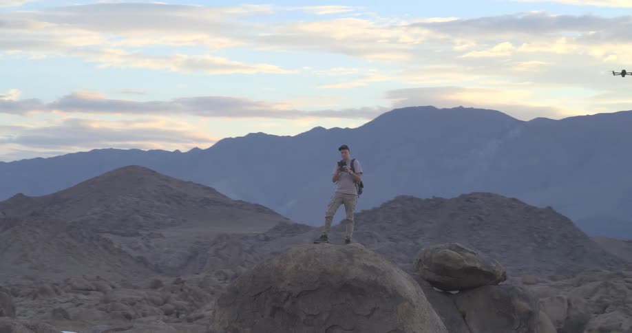 A drone pilot stands on a large boulder operating his drone in a vast rocky desert landscape at sunset