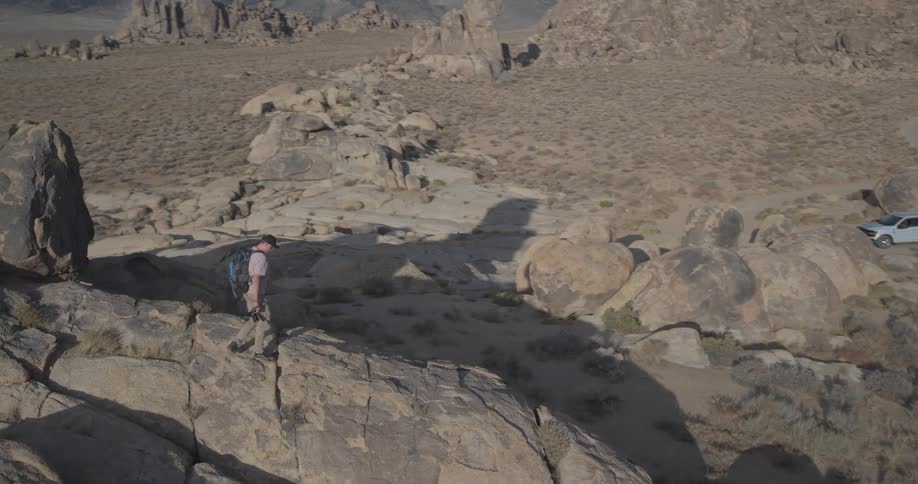 A lone hiker ascends a rocky formation in a vast desert landscape, revealing a stunning view of a mountain range under a blue sky