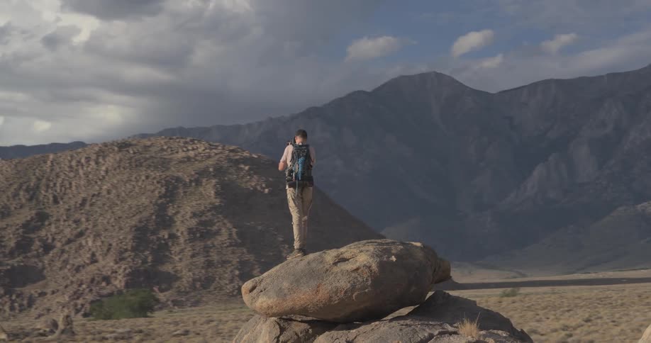 A male photographer with a backpack stands atop a large boulder in a desert, capturing the vast mountain scenery as the camera orbits around him