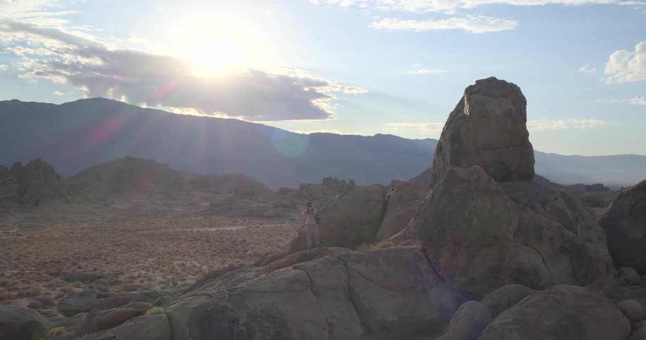 A photographer stands atop a large rock formation in the desert, capturing the expansive mountain landscape during a beautiful sunset