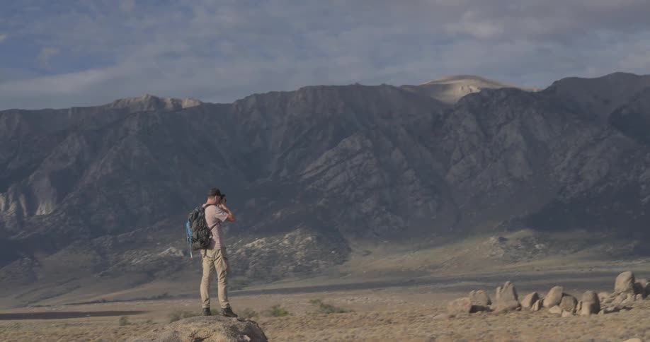 A male photographer stands on a large boulder and captures photos of the majestic mountain landscape before the camera pulls back to reveal the epic scenery