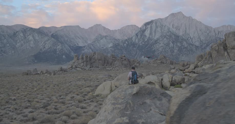 A drone pilot with a backpack sits on a boulder and flies a drone through a vast, scenic mountain valley at dusk