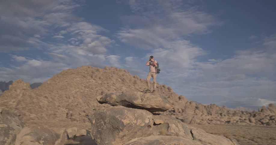 A male photographer with a backpack stands on a large boulder in a vast desert, capturing photos of the majestic mountain range