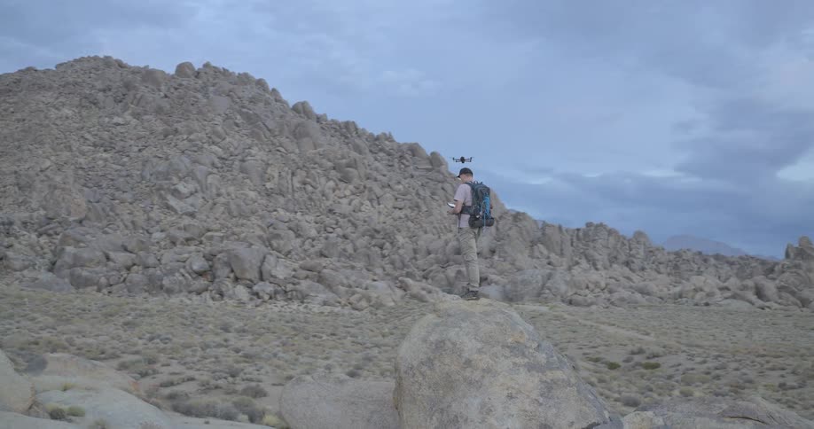 Slow-motion pan of a male adventurer standing on a large boulder in a vast desert landscape while flying a drone