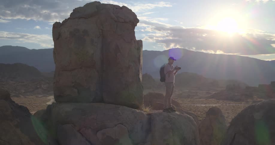 Aerial orbit of a photographer standing on a large rock formation taking pictures of the desert mountain landscape at sunrise