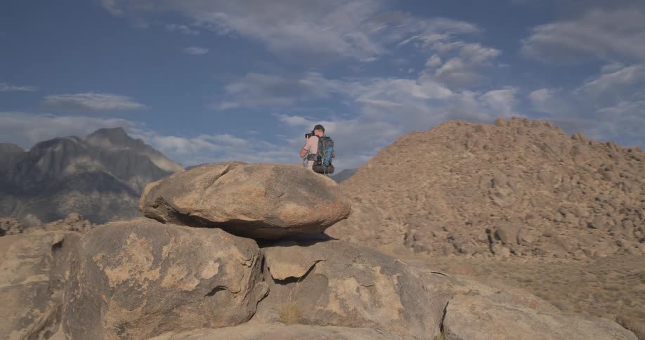 A male adventurer with a backpack climbs up a large boulder in a rocky desert and photographs the mountainous terrain