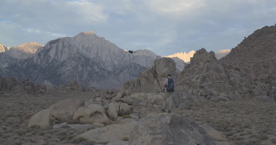 A drone pilot with a backpack launches a quadcopter in a rocky, arid landscape, revealing the stunning Sierra Nevada mountain range at sunrise