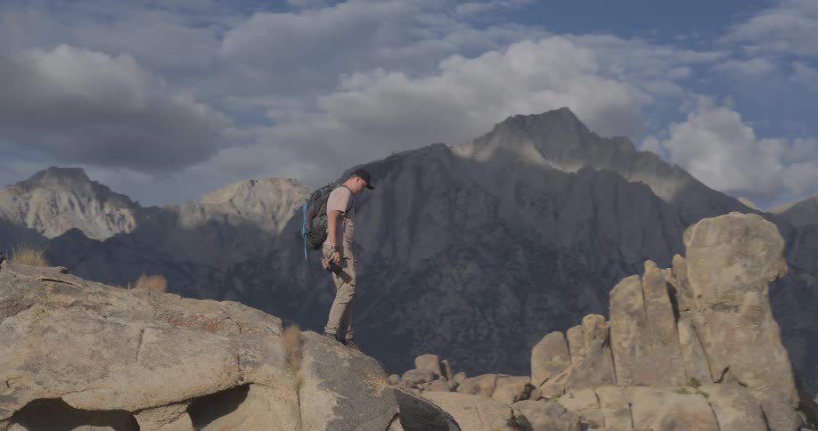 A male hiker with a backpack and camera carefully climbs over boulders in a vast mountain landscape in slow motion