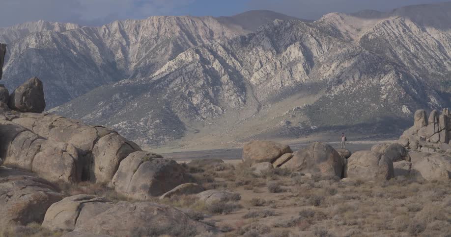 A lone hiker with a backpack stands atop a large boulder, gazing at the majestic Sierra Nevada mountains in the Alabama Hills