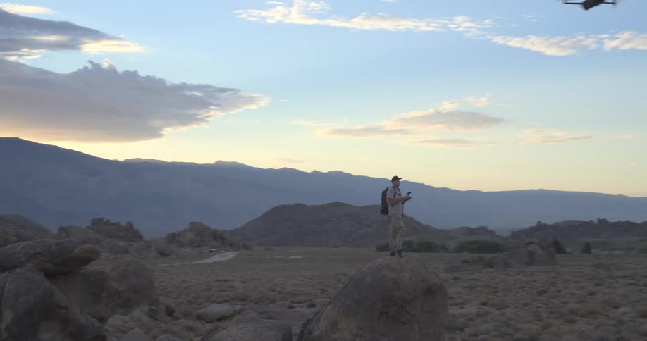 Orbiting shot reveals a hiker standing on a large boulder, operating a drone in a vast, rocky desert with distant mountains at sunset