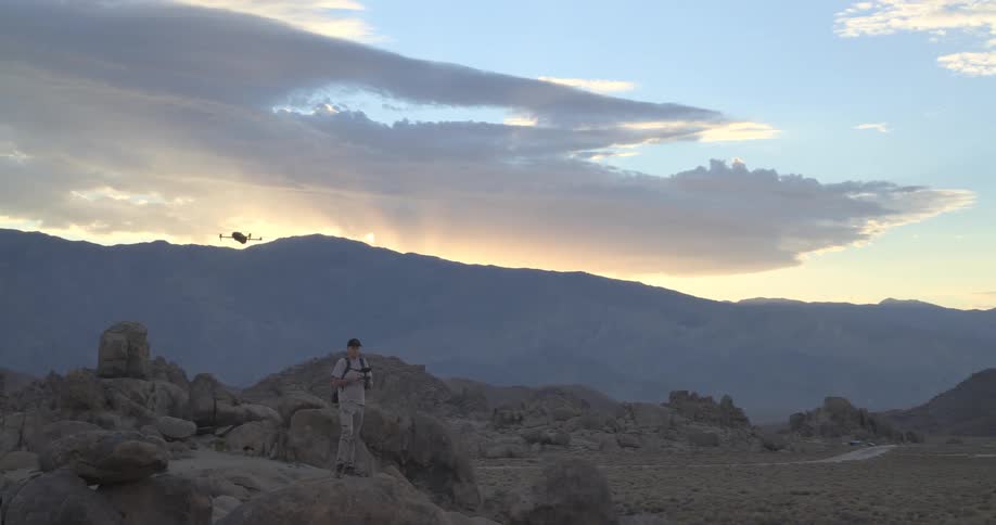 A drone pilot operates his aircraft from a rocky perch in a vast desert mountain landscape during a beautiful sunset