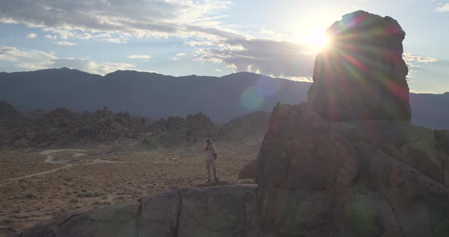 A photographer climbs a large rock formation in the desert to set up his camera and capture the beautiful landscape at golden hour