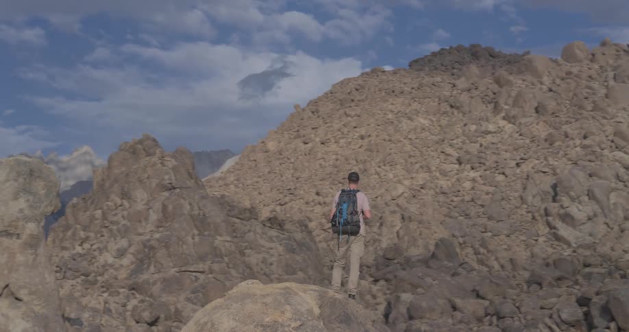 A male photographer with a backpack climbs a large boulder to take pictures of the majestic mountains in a vast, rocky desert landscape