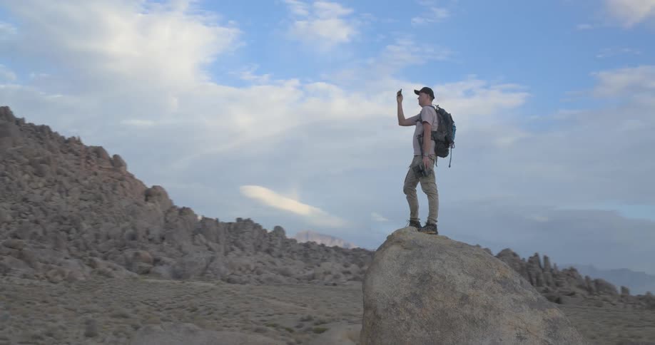 A lone hiker with a backpack stands on a boulder in a vast, rocky landscape, taking photos of the majestic mountain range with his phone at sunrise