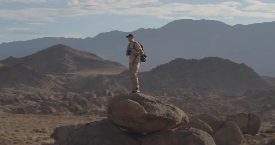 A photographer with a backpack stands on a boulder, capturing photos of the vast and rocky desert landscape during a hazy sunrise
