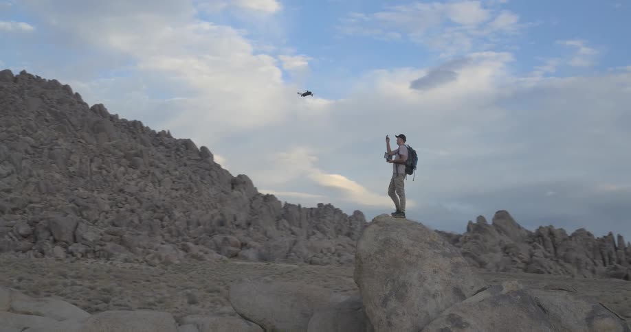 A hiker stands atop a large boulder, piloting a drone through a majestic desert mountain range during a beautiful sunrise