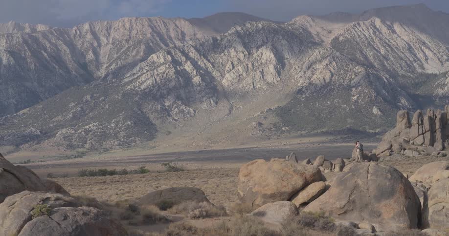 A lone hiker with a backpack stands atop a large boulder in a vast, rocky landscape, admiring the epic view of the Sierra Nevada mountain range