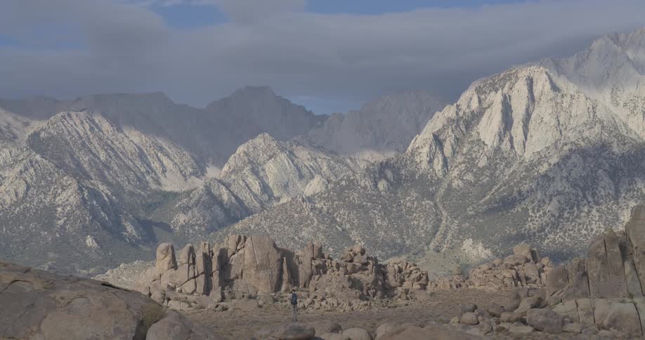 A lone hiker walks through the rocky Alabama Hills with the vast Sierra Nevada mountains looming in the background as the camera pulls back