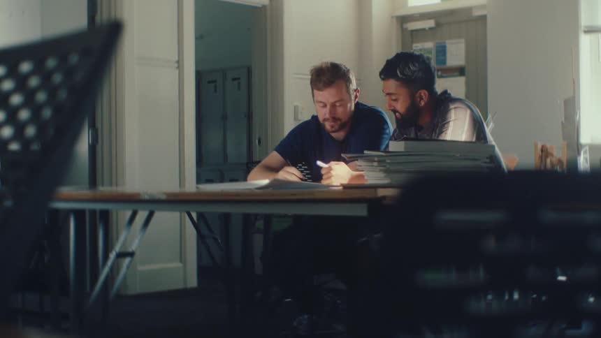 Two diverse male colleagues work together at a desk in a naturally lit office, focused on writing and discussing their project