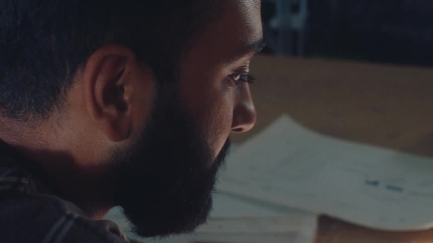 Close-up profile of a bearded man deeply focused on his work at a desk in a dimly lit room