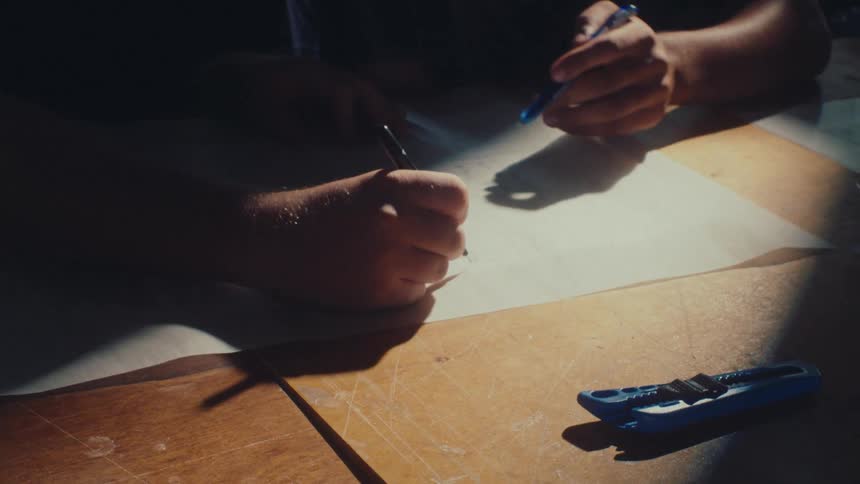 A close-up of two designers collaborating on a drawing, sketching on paper at a wooden workbench lit by a beam of light with dreamy vibe